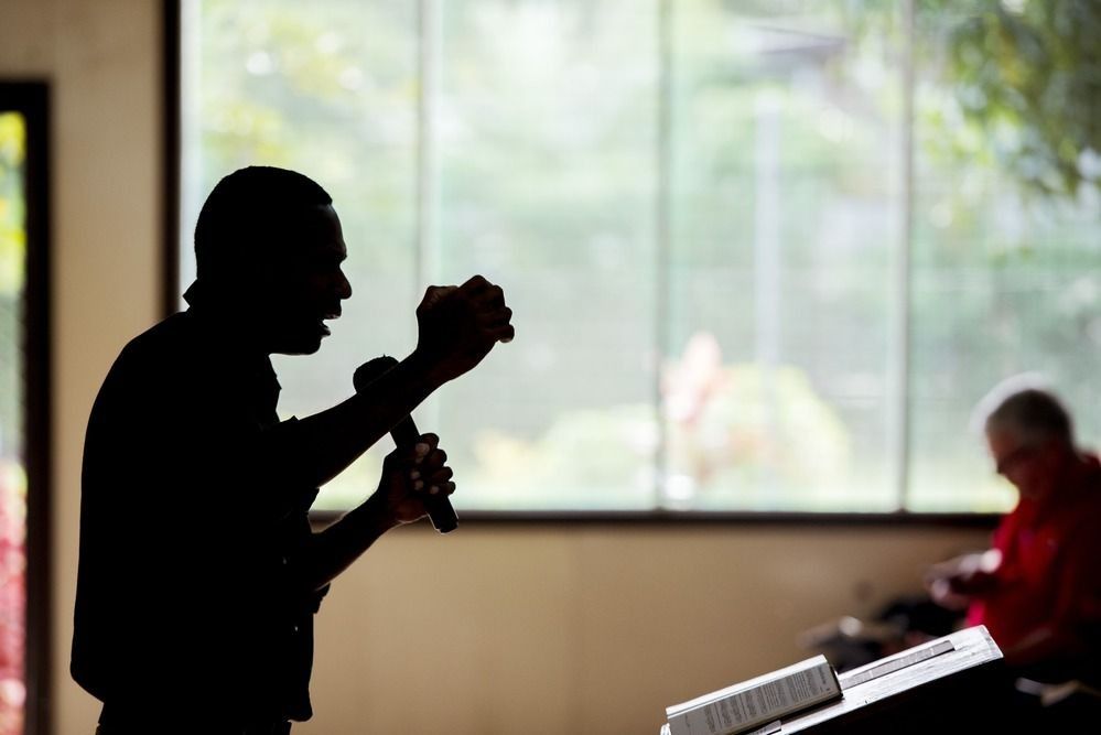 A silhouetted speaker gestures while holding a microphone at a podium, with a person seated in the background.