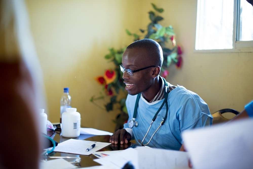 A healthcare provider in blue scrubs and a stethoscope sits at a desk with medical documents, speaking to a patient.