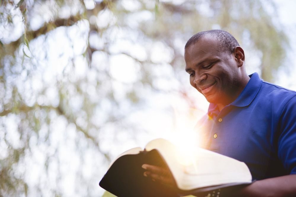 A smiling person in a blue polo shirt reads a book with sunlight glowing from the pages in an outdoor setting.