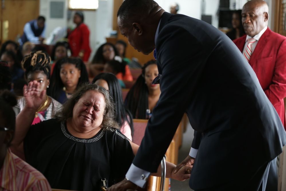 A person in a suit holds the hand of an emotional woman in a church setting while another person in a red jacket watches.