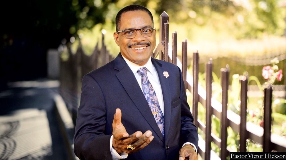 Pastor Victor Hickson in a dark suit and patterned tie, gesturing with one hand near a fence.