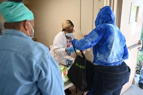 A masked medical professional in a white coat hands supplies to an individual wearing a blue hoodie and holding a tote bag.