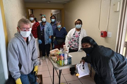 A group of people wearing face masks and gloves standing in a hallway behind a table containing food items.