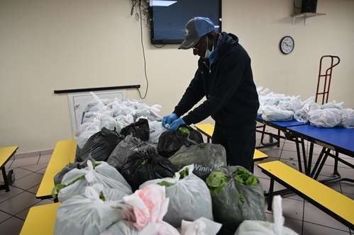 A person wearing a cap and blue gloves packs white and black plastic bags of food on tables in a bright, indoor space.