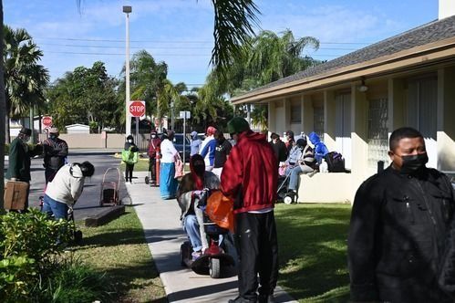 People wait in a line along a sidewalk on a sunny day outside a building, with some individuals using mobility scooters.