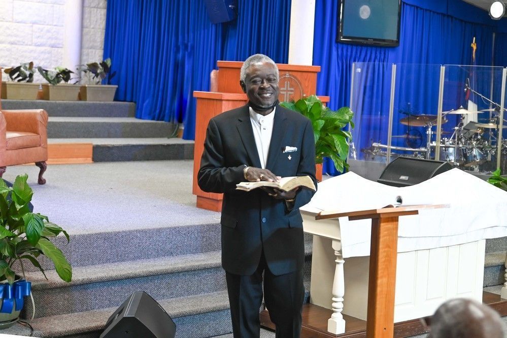 A smiling man in a suit holding an open book, standing on a church stage with blue curtains and a podium.