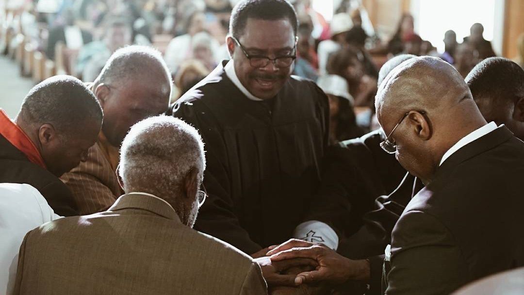A group of people standing in a circle with hands extended inward during a communal prayer service.
