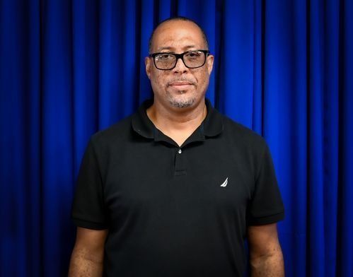 A person wearing glasses and a black polo shirt stands in front of a blue curtain backdrop.