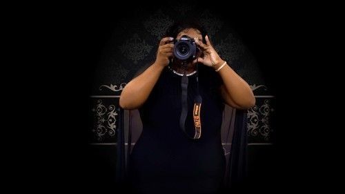 A person in a black outfit stands against a dark patterned background, holding a camera up to their face.