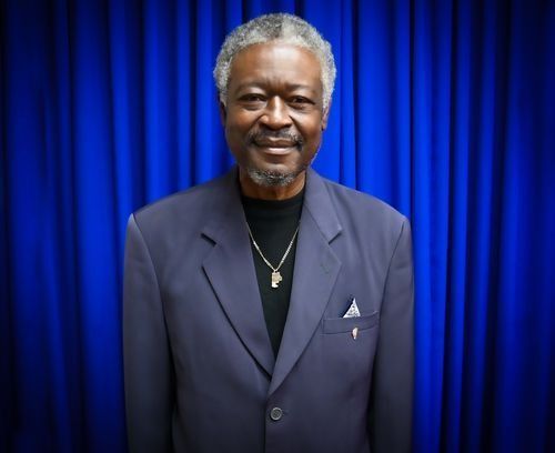 A person wearing a navy blazer and black shirt with a gold chain, standing in front of a blue pleated curtain.
