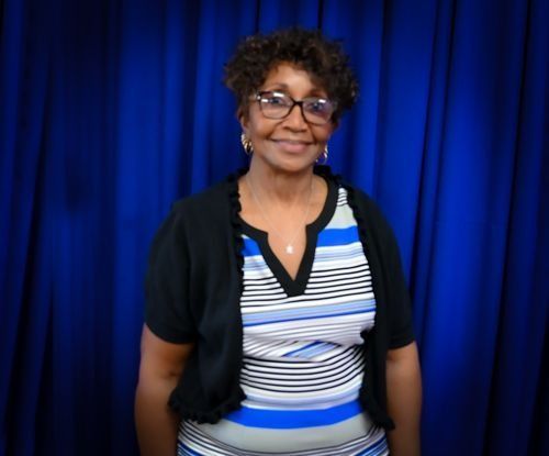A person wearing glasses and a striped shirt with a black cardigan stands in front of a blue curtain backdrop.