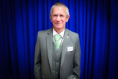 A man in a grey three-piece suit and a striped green tie standing in front of a blue curtain backdrop.