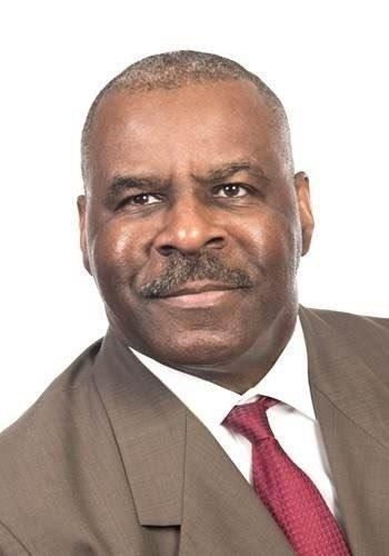 A man wearing a tan suit, white shirt, and maroon necktie, looking directly at the camera with a neutral expression.