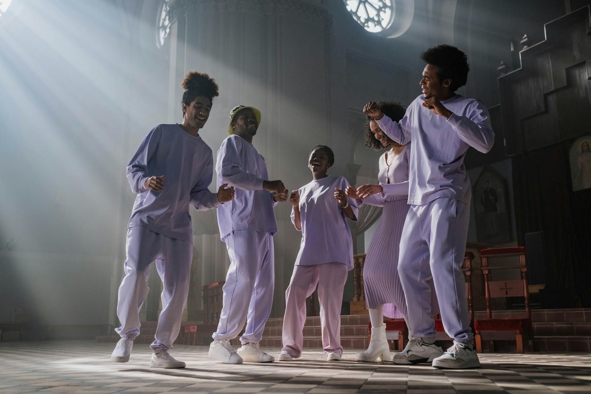 Five people in matching light-colored outfits dance joyfully in a sunlit, historic building with high ceilings.
