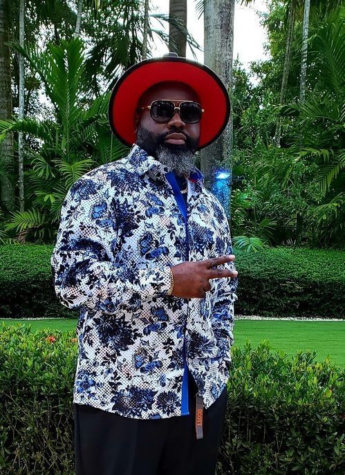 A man in a floral shirt, black pants, and a red-brimmed fedora gives a peace sign in a lush, green tropical garden.