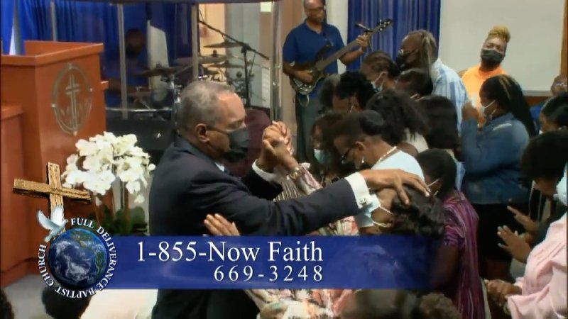 A man in a suit leads a prayer over a group in a church, with a contact phone number overlaid on the bottom of the screen.