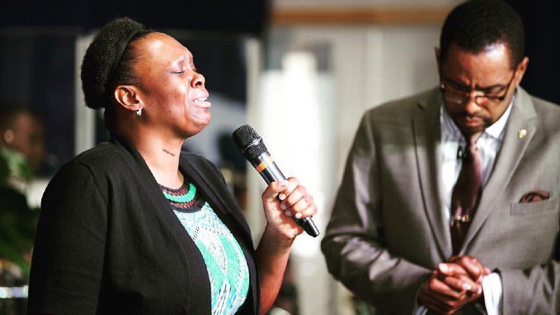 A person singing into a microphone with eyes closed, while another person stands nearby with hands clasped in prayer.