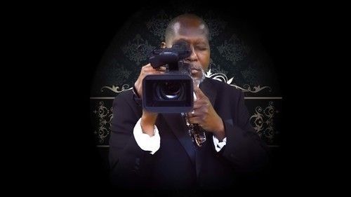 A person in a dark suit holds a professional video camera while looking through the viewfinder against a dark background.