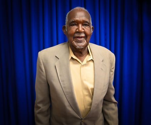 A smiling man wearing a beige blazer and button-down shirt stands in front of a blue curtain.