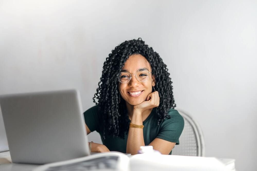 A person with dark curly hair and glasses smiles while leaning their chin on their hand, seated at a desk with a laptop.