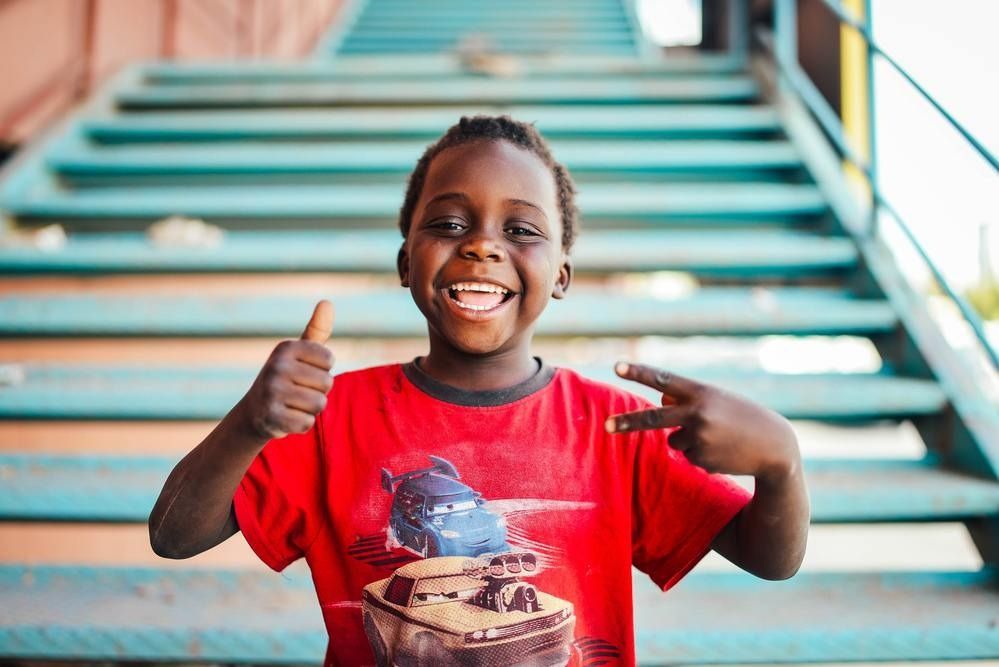 A smiling child in a red t-shirt makes a thumbs-up and a peace sign while sitting in front of teal metal stairs.