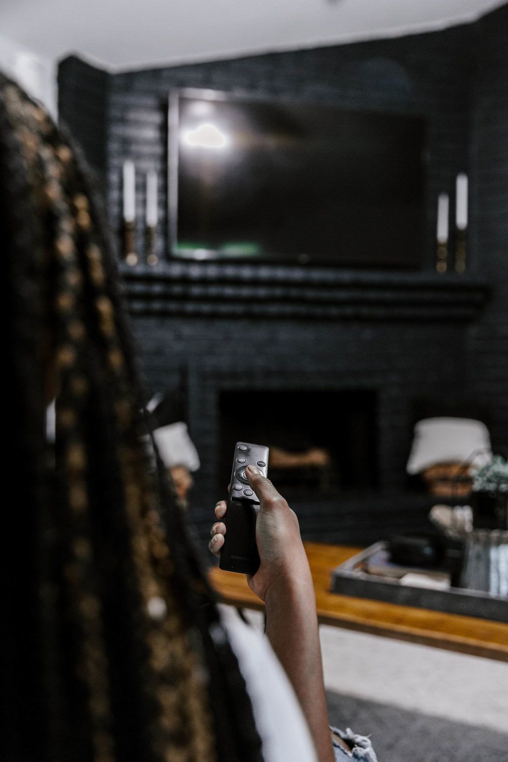 A person holding a remote in a dark, cozy living room with a brick fireplace and wall-mounted television.