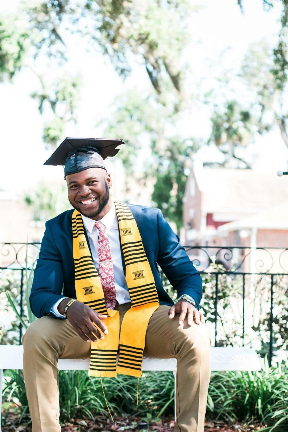 A graduate in a blue blazer, khaki pants, and cap sits smiling, wearing a yellow stole with black patterns outdoors.