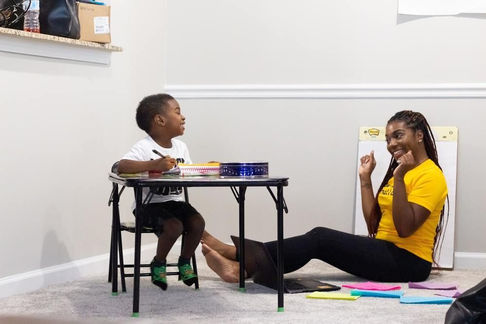 A person in a yellow shirt sits on the floor across from a child at a small table, both smiling during an activity.