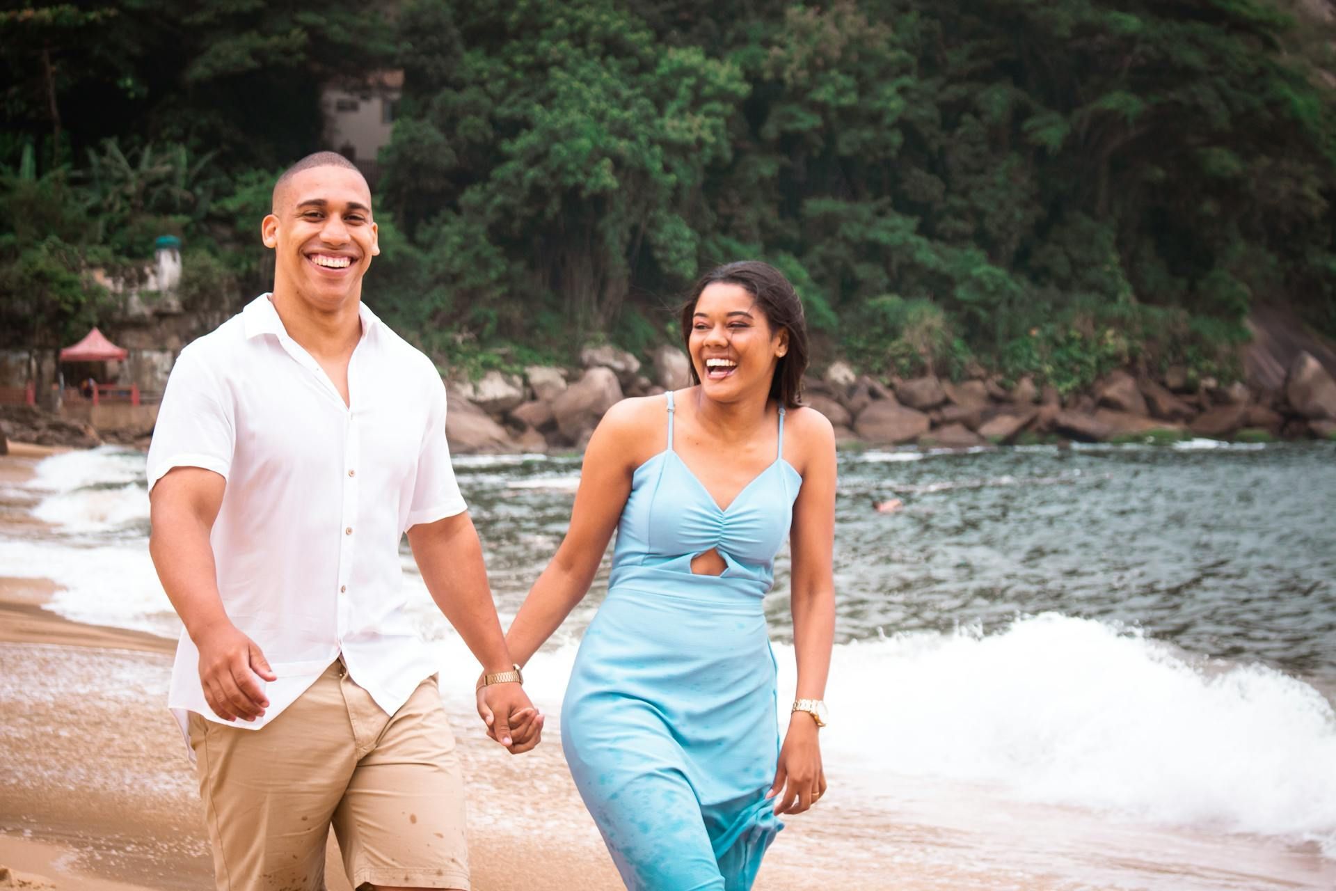A couple in casual clothing holds hands and laughs while walking along a sandy beach by the ocean.