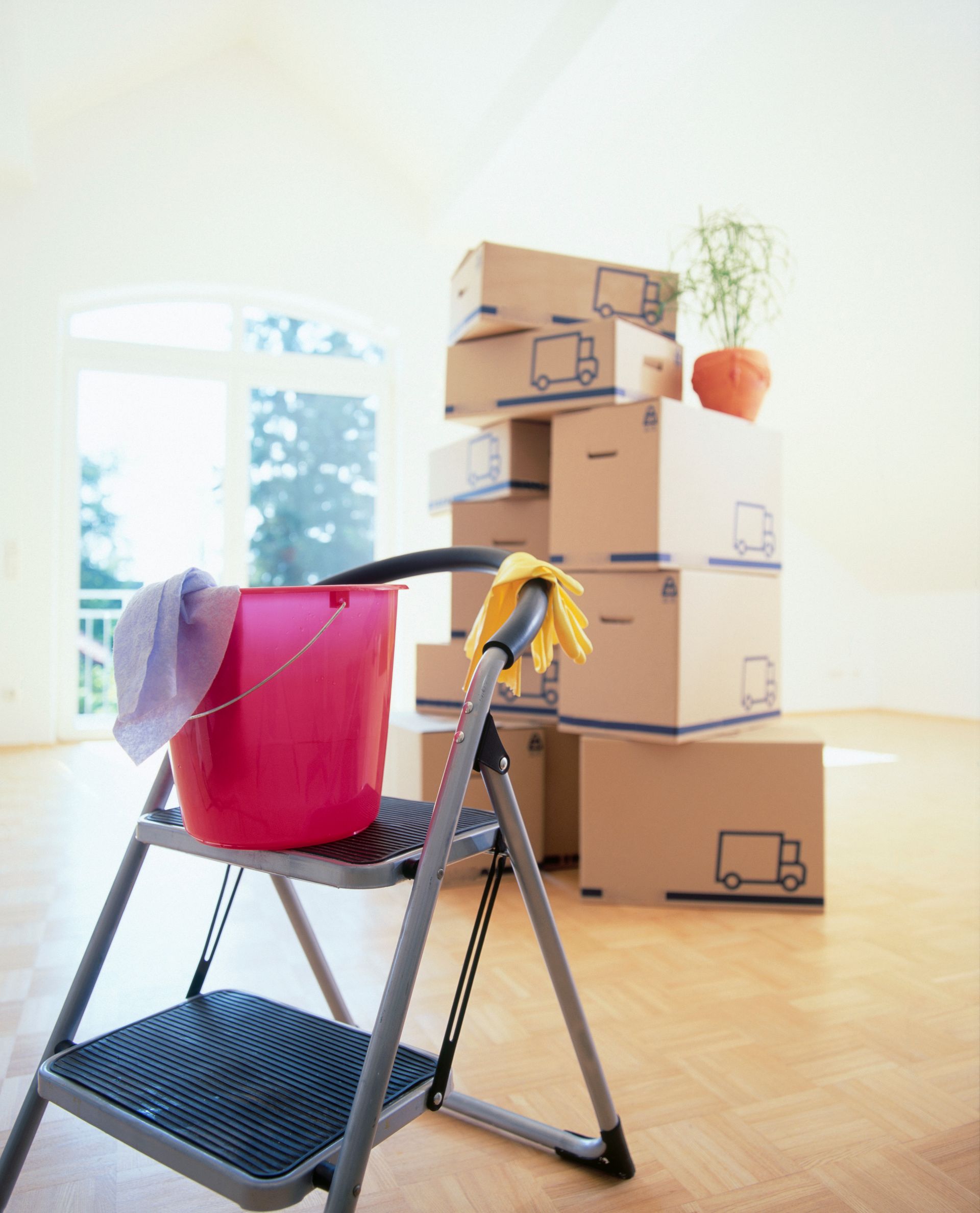 Stacked moving boxes beside a black folding step stool with a red bucket and yellow gloves in a bright room