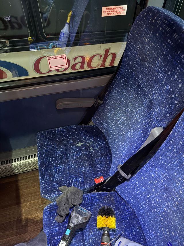 Blue speckled bus seat beside a window with a black seatbelt and a small yellow-black object on the cushion