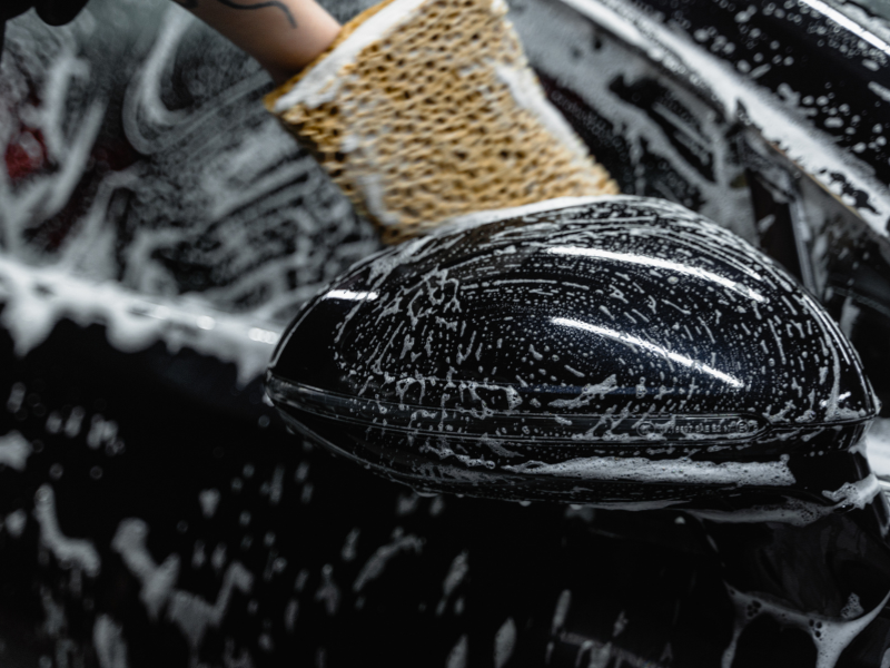 Black car being washed, covered in suds with a sponge scrubbing the roof Close-up of a black car being washed with soap suds and a sponge