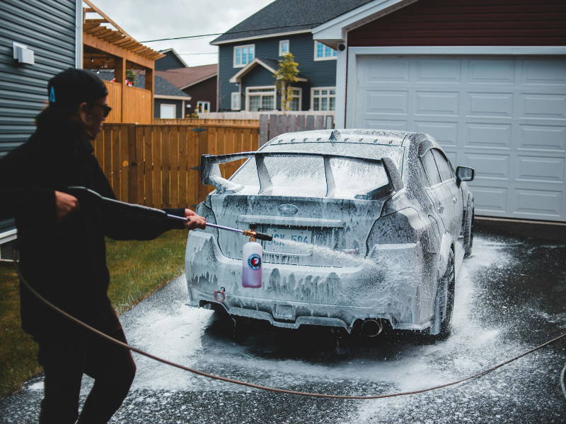 Person pressure-washing a foam-covered car in a driveway beside a garage
