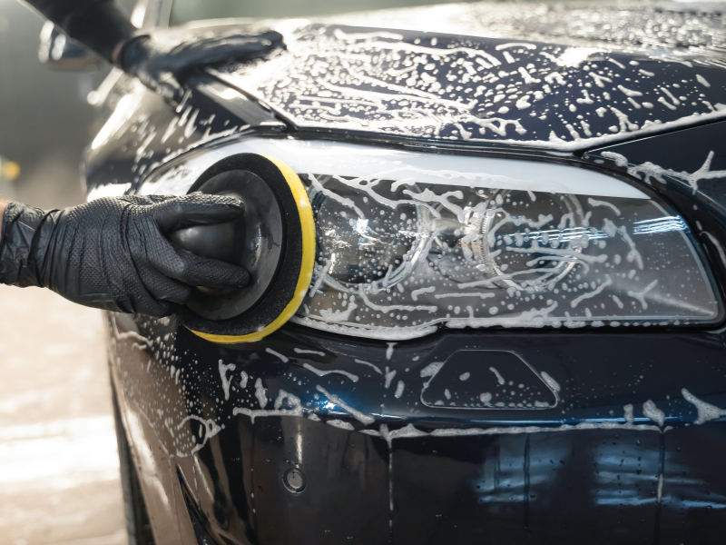 Gloved hand polishing a black car headlight with soapy foam on the hood