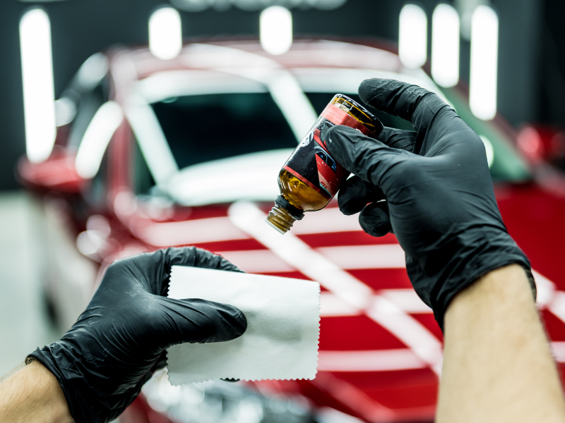 Gloved hands applying car polish to a red vehicle with a cloth in a detailing shop
