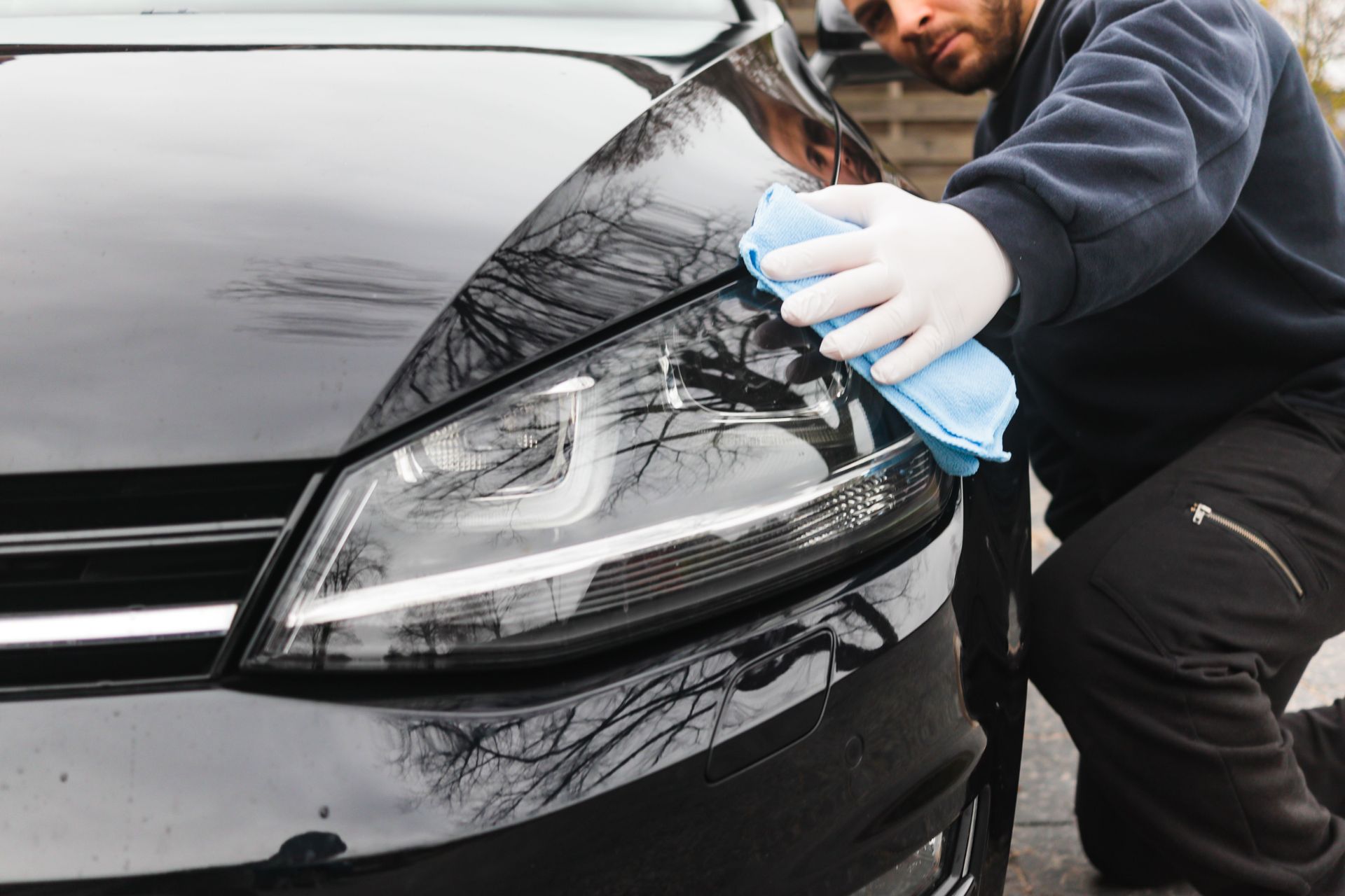 Person cleaning a black car’s headlight with a blue cloth and spray bottle