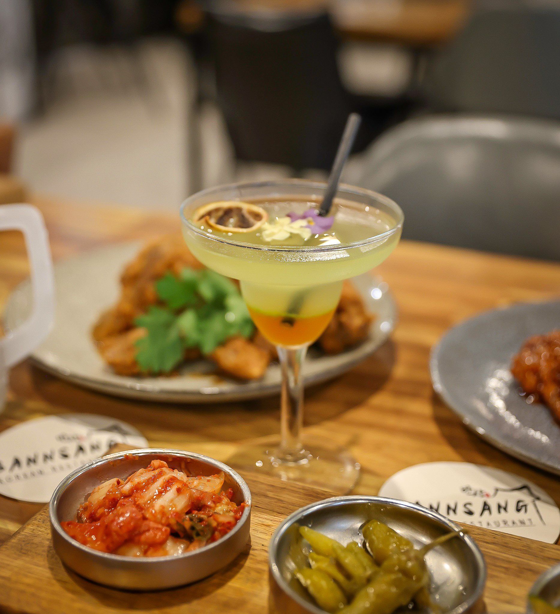Cocktail with a garnish, fried food, and small dishes of kimchi and pickles on a wooden table.