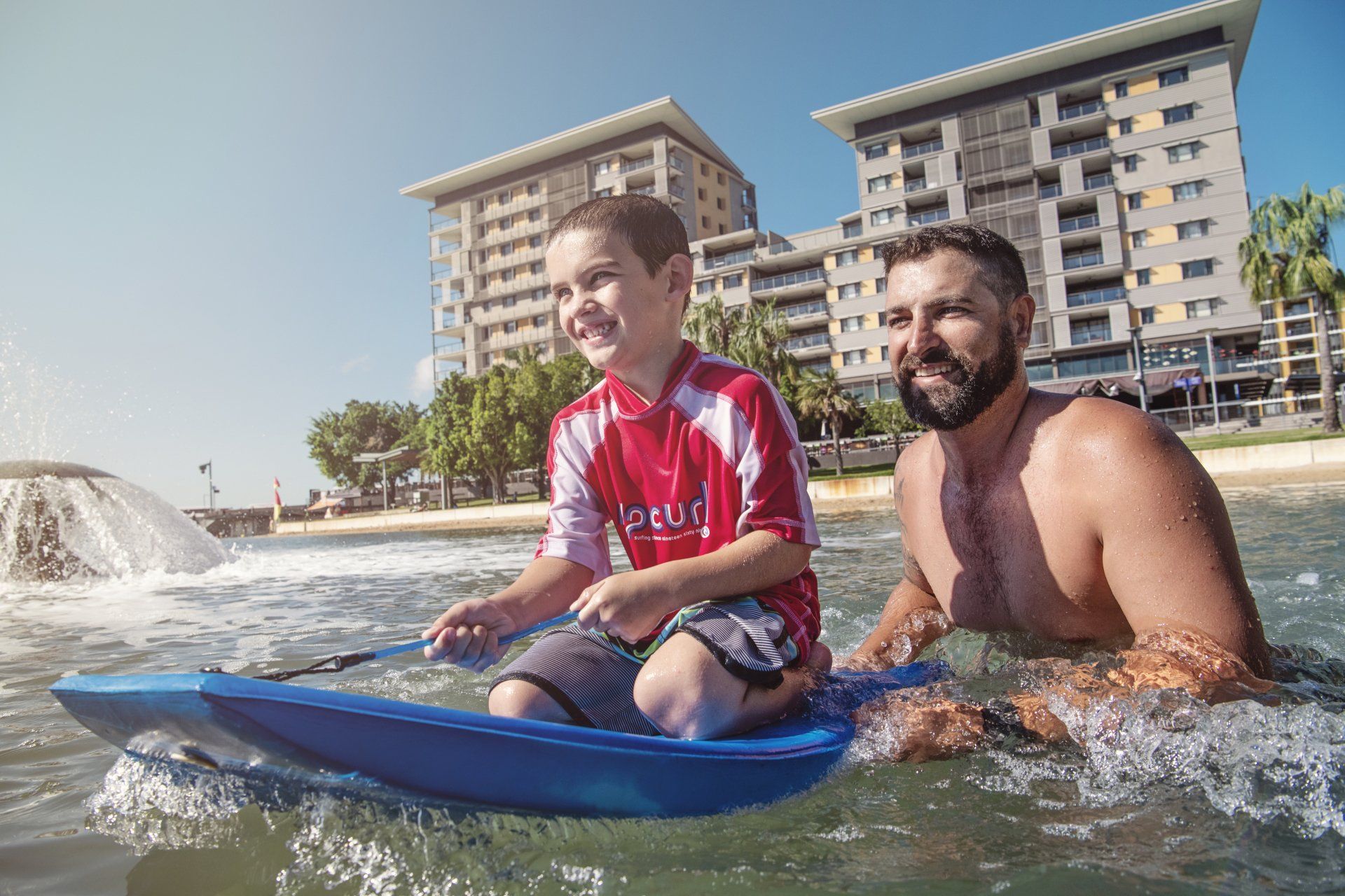 Boy and man ride a blue water sled in a shallow body of water near a beach and buildings.