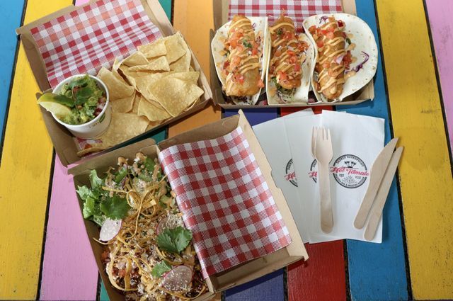 Mexican food spread: tacos, chips, guacamole, noodles in brown cardboard containers on a colorful table.