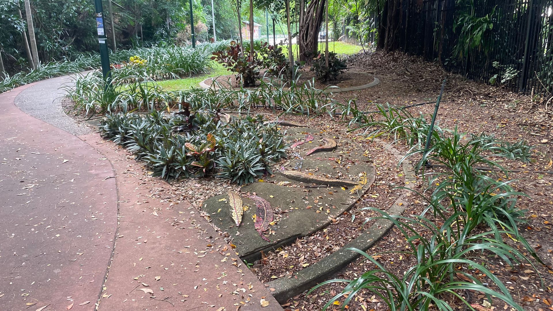A paved path curves past garden beds with plants and fallen leaves; trees in background.