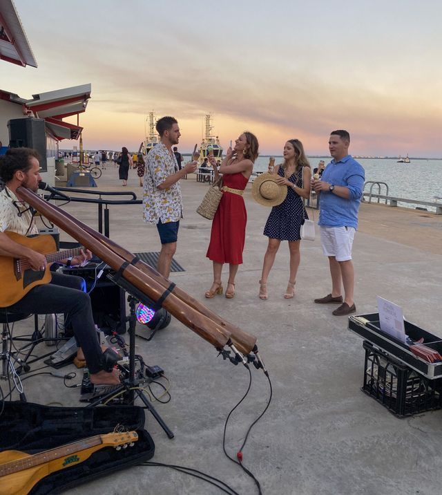 Musician plays guitar and didgeridoo for a group on a pier at sunset. People smile, enjoying the music.