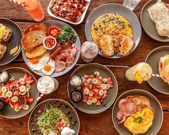 Breakfast spread on a wooden table: eggs, bacon, toast, waffles, pancakes, omelets, fruit, drinks.