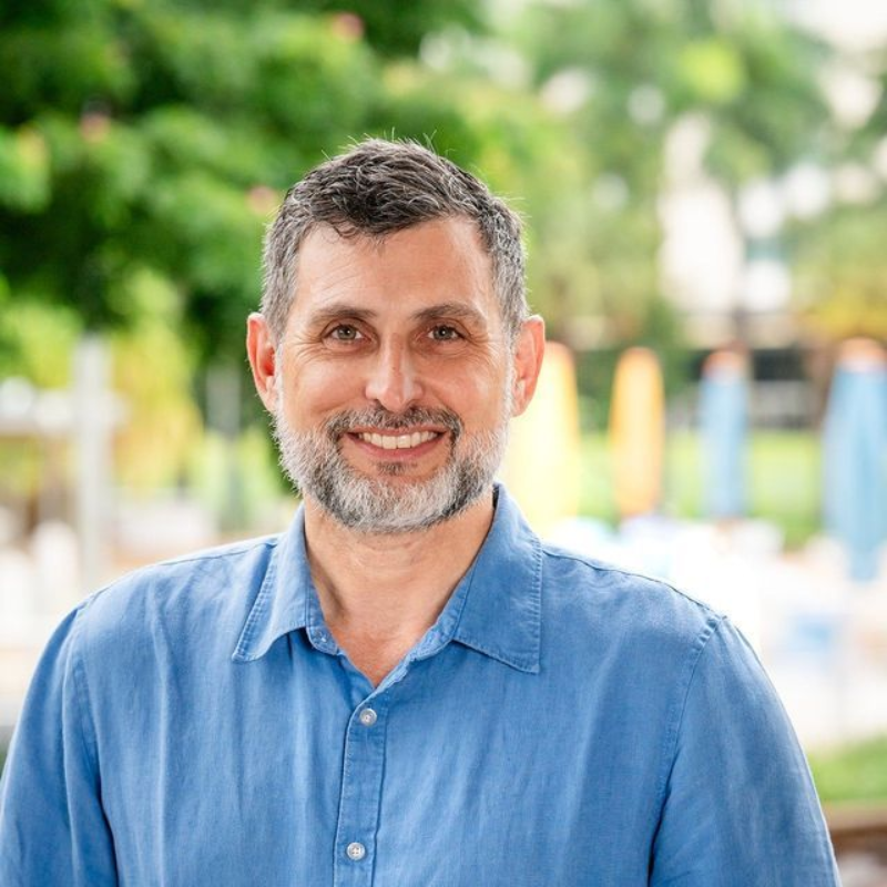 Man with a gray beard smiles, wearing a blue shirt, in a blurred outdoor setting.