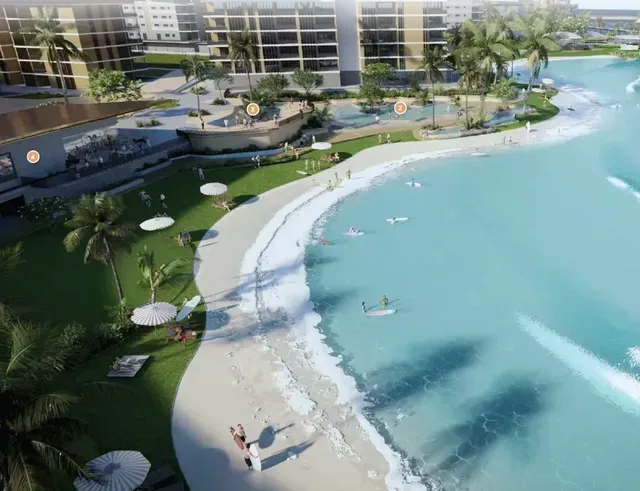 Aerial view of a wave pool beach with surfers, buildings, and palm trees. Light blue water, green grass, and sunny day.