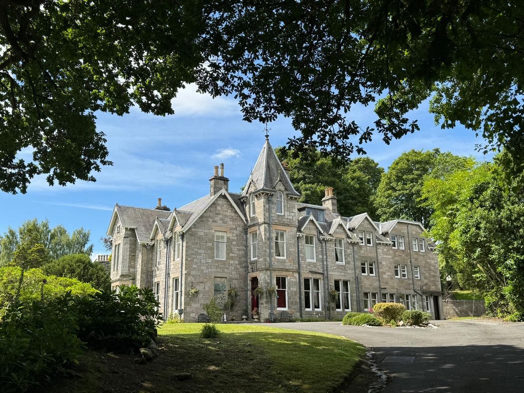 Stone manor house with gabled roofs, surrounded by trees and greenery under a clear blue sky