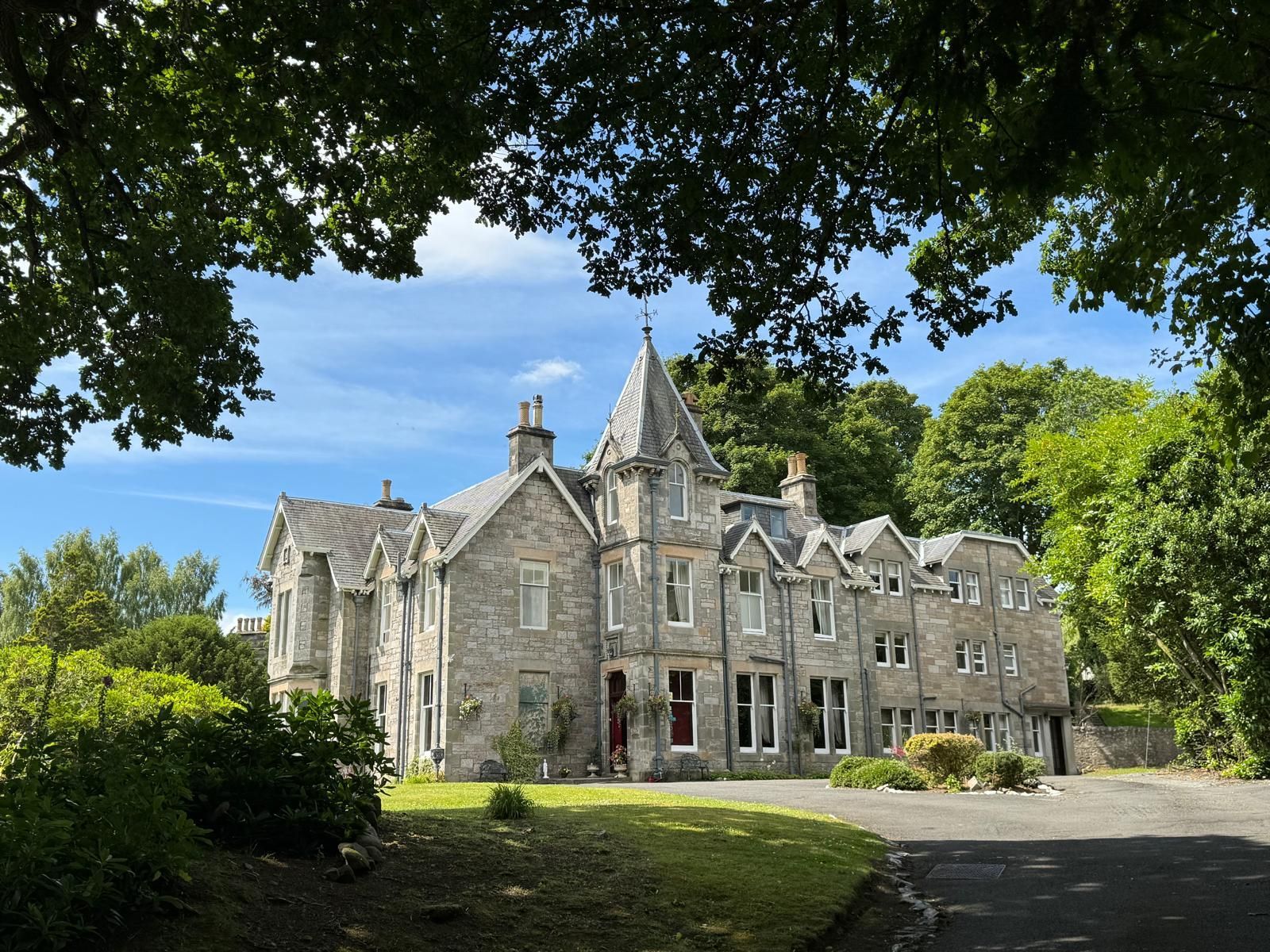 Stone manor house with gabled roofs, surrounded by trees and greenery under a clear blue sky