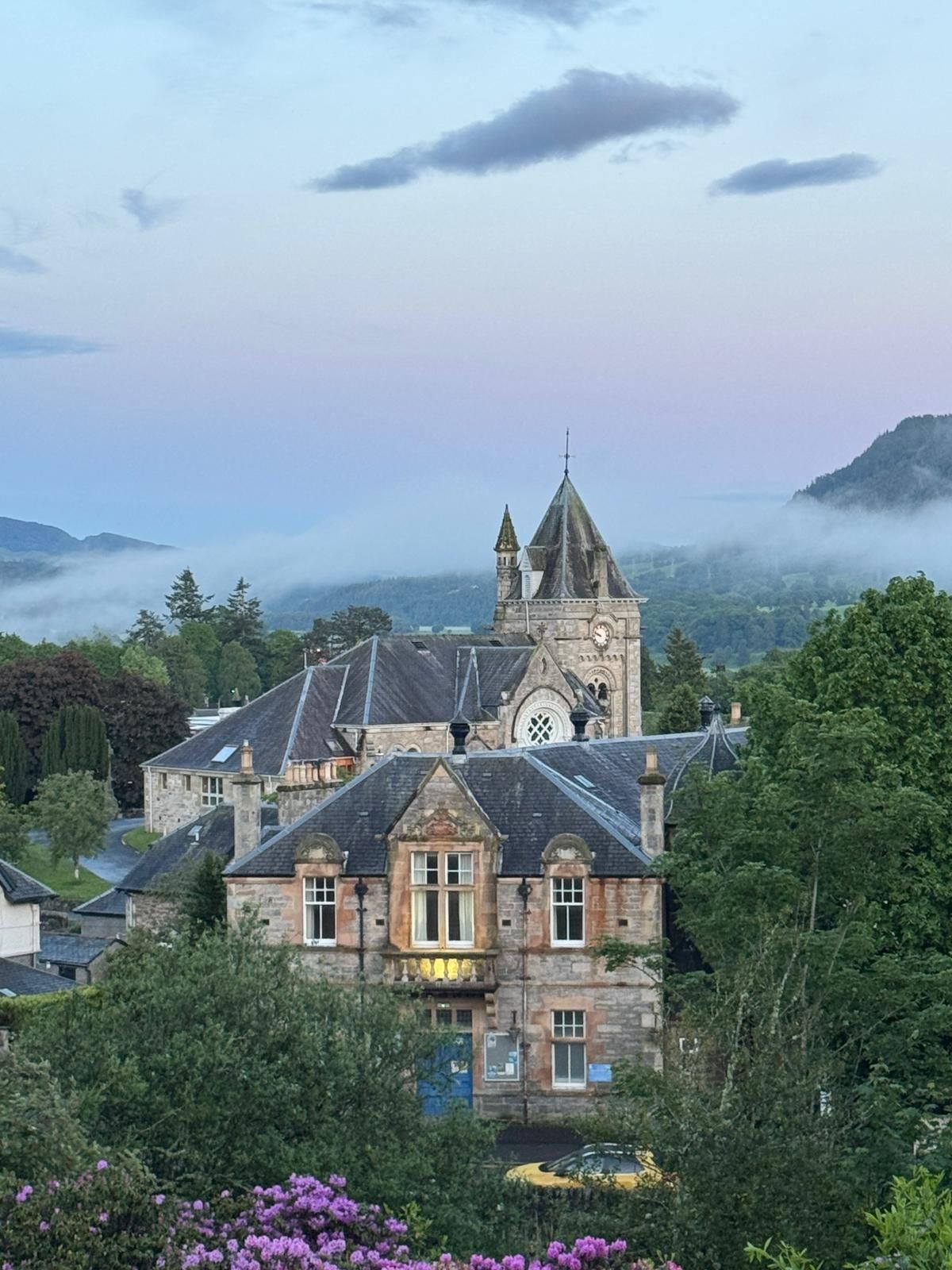 Stone chateau with lit windows and twin towers amid misty mountains and purple flowers at dusk