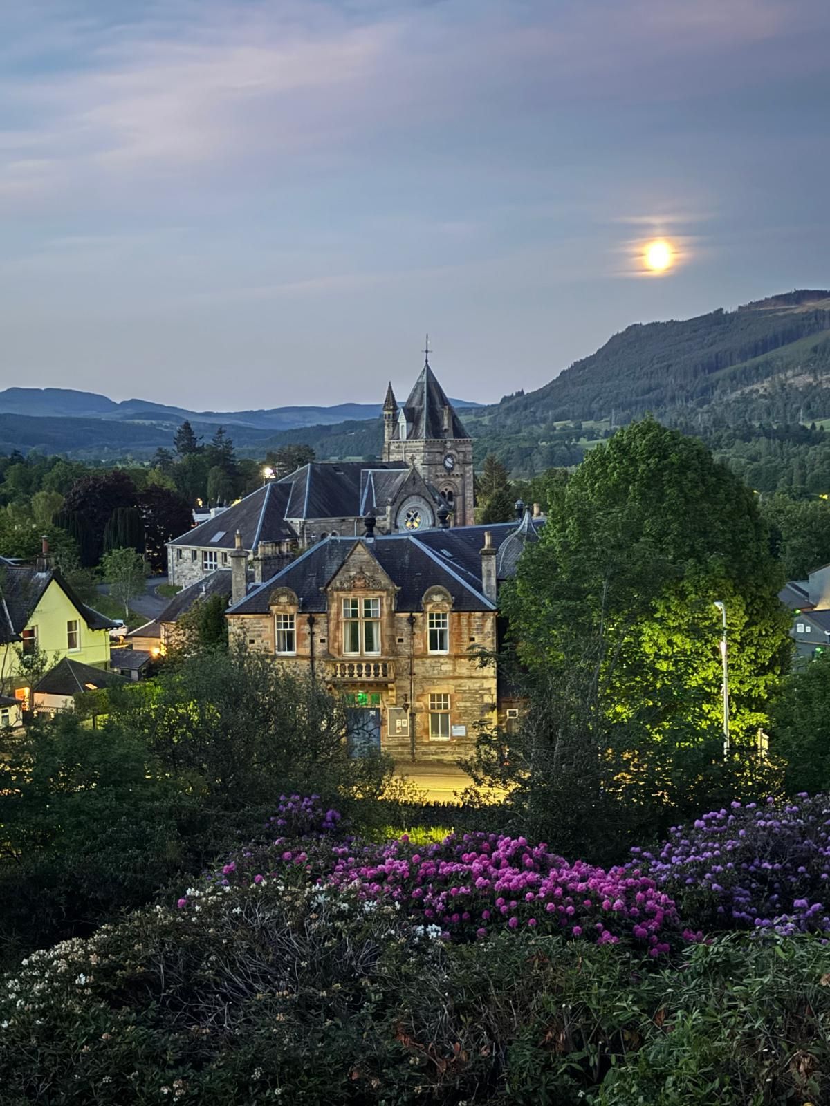 Lit stone castle lit at dusk beside a moonlit hillside, with flowers and trees in the foreground