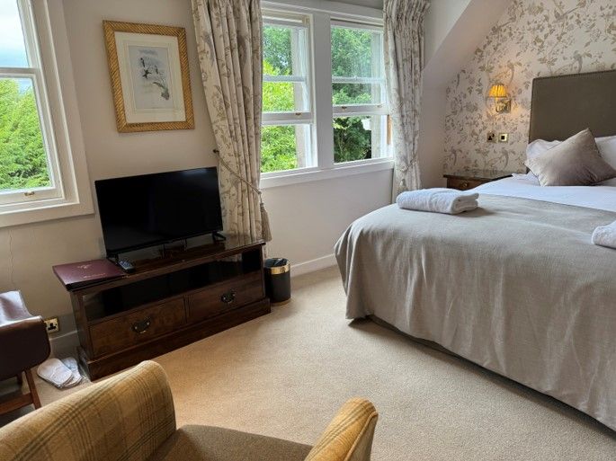 Bright bedroom with large windows, a bed, TV on a wooden dresser, and a wicker chair near the foreground