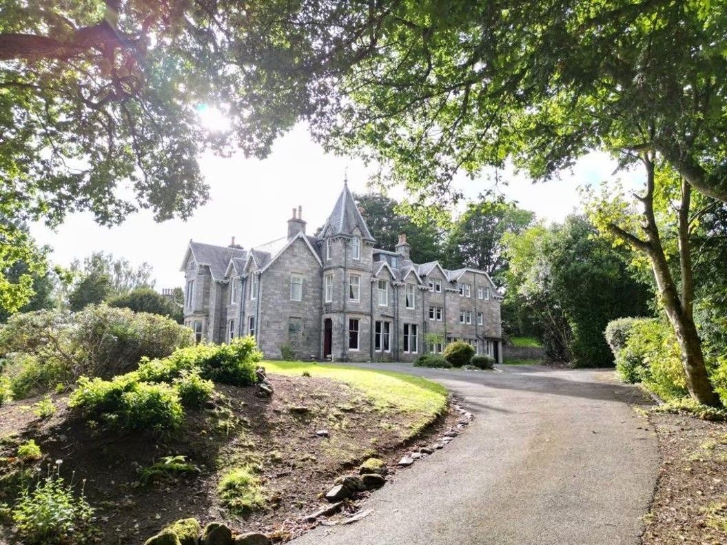 Stone manor house on a curving driveway, framed by lush green trees and shrubs in bright daylight.